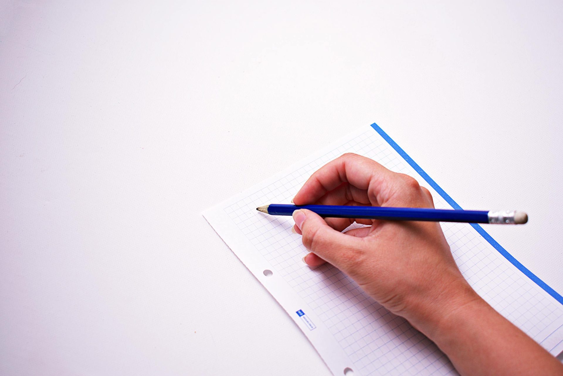 Close-up of a hand writing on graph paper using a blue pencil, on a white background.